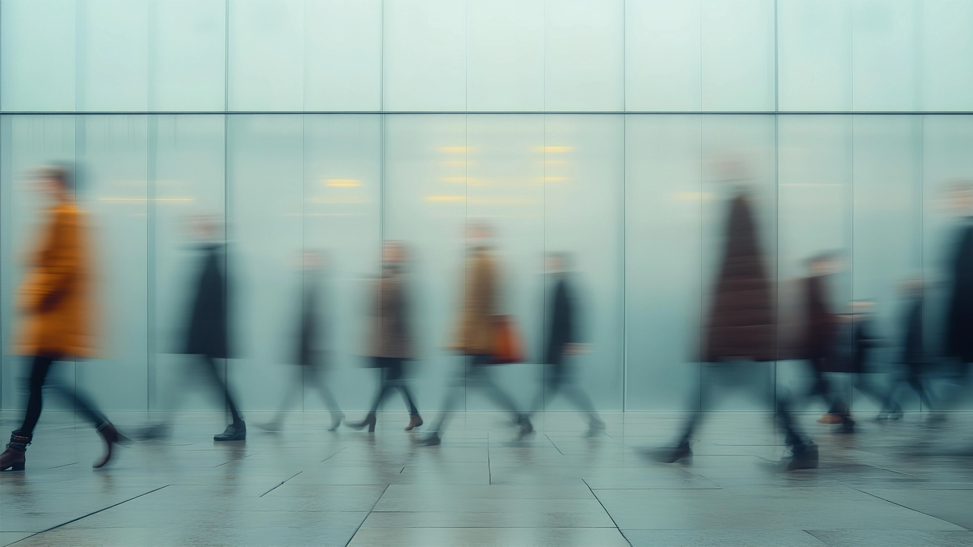 Verschwommene Menschenmengen in Bewegung vor einer Glasfassade. Die Personen sind unscharf dargestellt, sodass Bewegung und Hektik des urbanen Alltags betont werden. Die Farbtöne sind überwiegend kühl, mit einzelnen Farbakzenten durch Mäntel in Braun-, Orange- und Beigetönen.